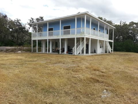 Cut field in front of a blue two-story stilt house with a wraparound porch and moss-draped oaks behind it