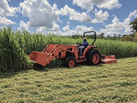 Orange Kubota tractor with a rotary cutter bush-hogging tall grass, leaving a cut path beside the uncut field